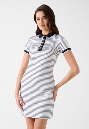 Młoda kobieta nosząca białą sukienkę z krótkim rękawem, z cienkimi czarnymi poziomymi paskami i granatowym kołnierzykiem z guzikami.