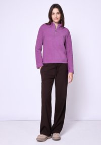 Pullover in pile con zip a un quarto di colore viola, abbinato a pantaloni ampi a righe marroni scure. Calzature in colori neutri con dettagli testurizzati.