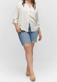 Lightweight, cream button-up shirt with rolled sleeves paired with distressed denim shorts. Model wears yellow sandals and stands against a neutral background.