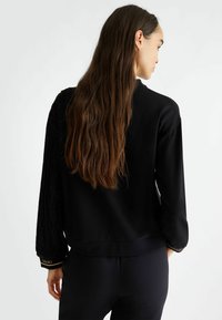 Maglione nero con manica sinistra texturizzata, corpo liscio, orlo a coste e polsini con dettagli dorati. Presenta un fit rilassato e maniche lunghe.