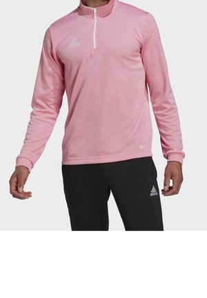 Veste de sport rose clair avec un col zippé à moitié, des manches longues, des poignets côtelés et des détails subtils du logo Adidas. Portée avec un pantalon noir.