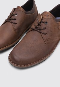 Chaussures en cuir marron décontractées pour hommes, à lacets, avec une finition texturée, des détails de couture et des semelles en caoutchouc sur un fond gris clair.