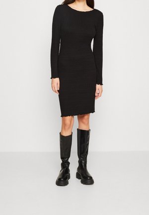 Schwarzes langärmeliges Kleid aus strukturiertem Stoff, figurbetonter Schnitt und gewelltem Saum. Kombiniert mit knielangen schwarzen glänzenden Stiefeln.