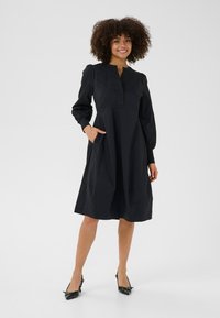 Moteris stovi ir šypsosi, dėvėdama ilgomis rankovėmis juodą midi ilgumo suknelę su kišenėmis ir juodus mažo kulno batus, fonas – paprastas baltas.