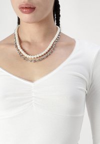 Collana a strati con perle bianche e un elemento in catena d'argento, indossata sopra una maglietta bianca attillata con scollo a V e dettagli arricciati.