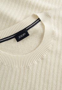 Cremefarbener Pullover mit strukturiertem Fischgrätmuster, geripptem Kragen und schwarzen sowie braunen Streifenakzenten im Inneren des Halsausschnitts. Markenetikett sichtbar.