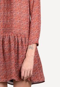 Röd blommig klänning med långa ärmar, samlad vid midjan, med en texturerad tyg och en utställd kjol. Inkluderar synlig tatuering på handleden.