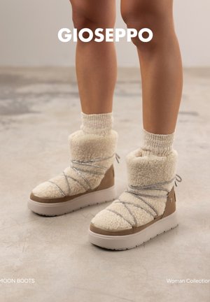 Kobieta nosząca kremowe buty moon boots wyściełane polarem, ze srebrnymi sznurówkami i grubymi białymi podeszwami, w połączeniu z pasującymi wełnianymi skarpetami, stojąca na betonowej podłodze.