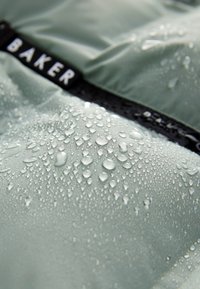 Waterdruppels bedekken groene stof met een zwarte strook met het label "BAKER", die de waterbestendige materiaaltextuur benadrukt.