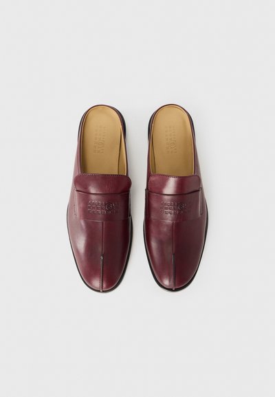 Ett par burgunderfärgade läderslip-on loafers med svarta präglade siffror på ovansidan, tanfärgade innersulor och runda tår mot en vit bakgrund.