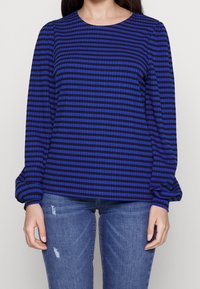 Blue and black striped long-sleeve top with a rounded neckline and gathered sleeves, made of a textured fabric. Paired with distressed blue jeans.