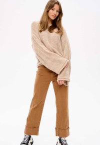 Beige Strickpullover mit weiten Ärmeln; kombiniert mit hellbraunen, schmal zulaufenden Hosen; mit aufgerollten Bündchen und schwarz-weißen Sneakern.