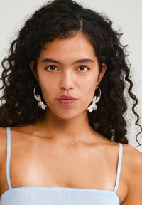 Silver hoop earrings with floral accents, featuring a textured design. Model wears a light blue spaghetti strap top. Skin tone is neutral.