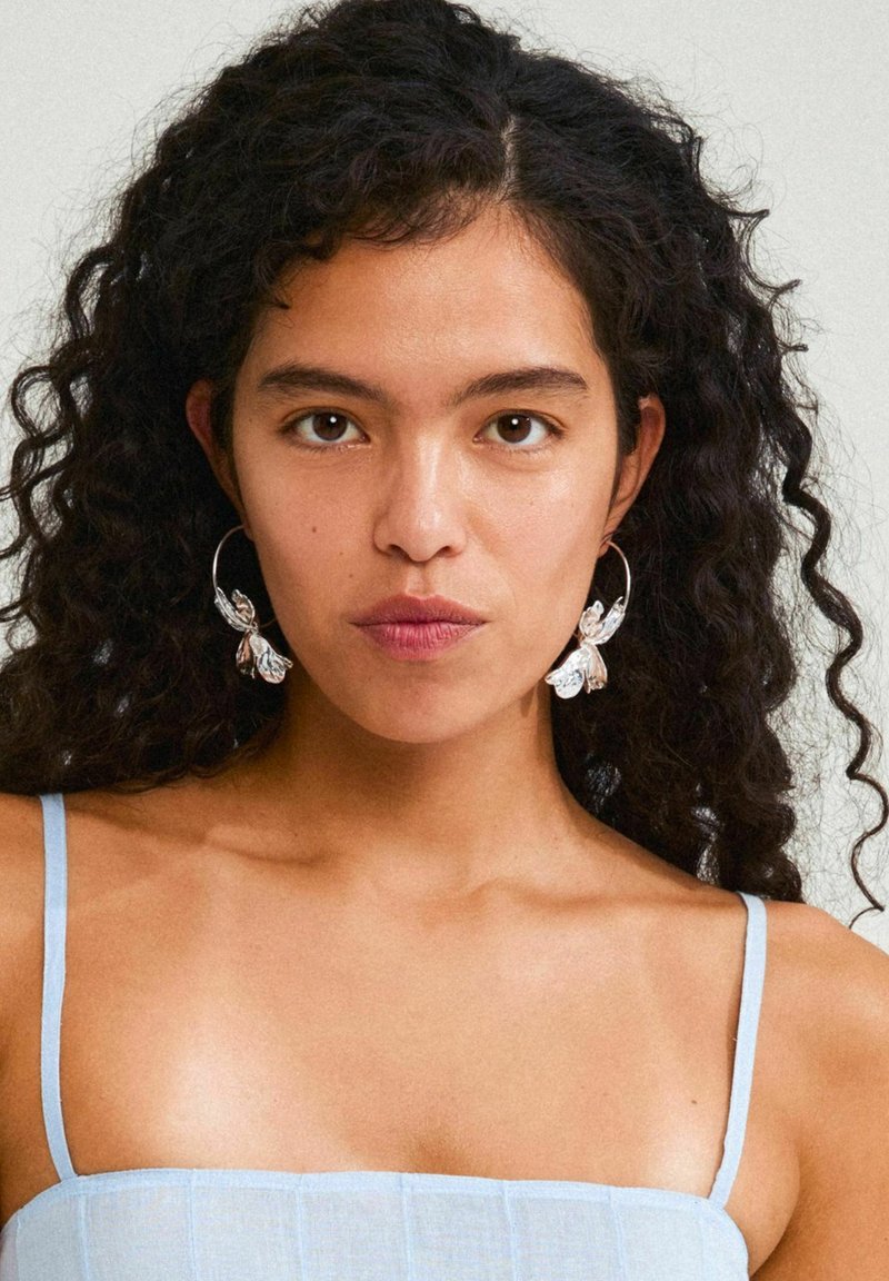Silver hoop earrings with floral accents, featuring a textured design. Model wears a light blue spaghetti strap top. Skin tone is neutral.
