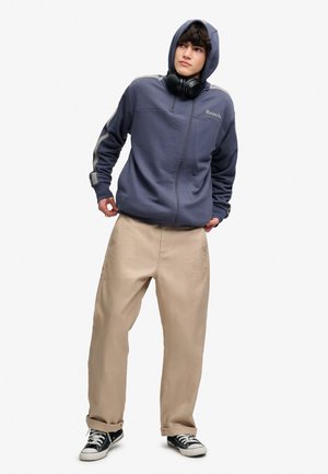 Ung mand iført en blå hættetrøje, beige bukser, sorte sneakers og sorte høretelefoner om halsen, stående foran en hvid baggrund.