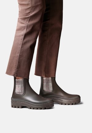 Personne portant un pantalon marron court et des bottes en caoutchouc épaisses marron foncé avec des panneaux latéraux élastiques à motifs.