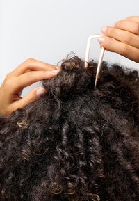 Kädet kiinnittävät kiharaiset hiukset bun-malliin suuren U-muotoisen kultaisen hiusneulan avulla vaaleaa taustaa vasten.