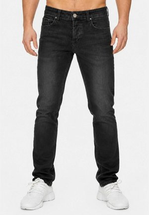 Man draagt slim-fit zwarte jeans en witte sneakers, staand met ontspannen handen langs het lichaam tegen een effen achtergrond.