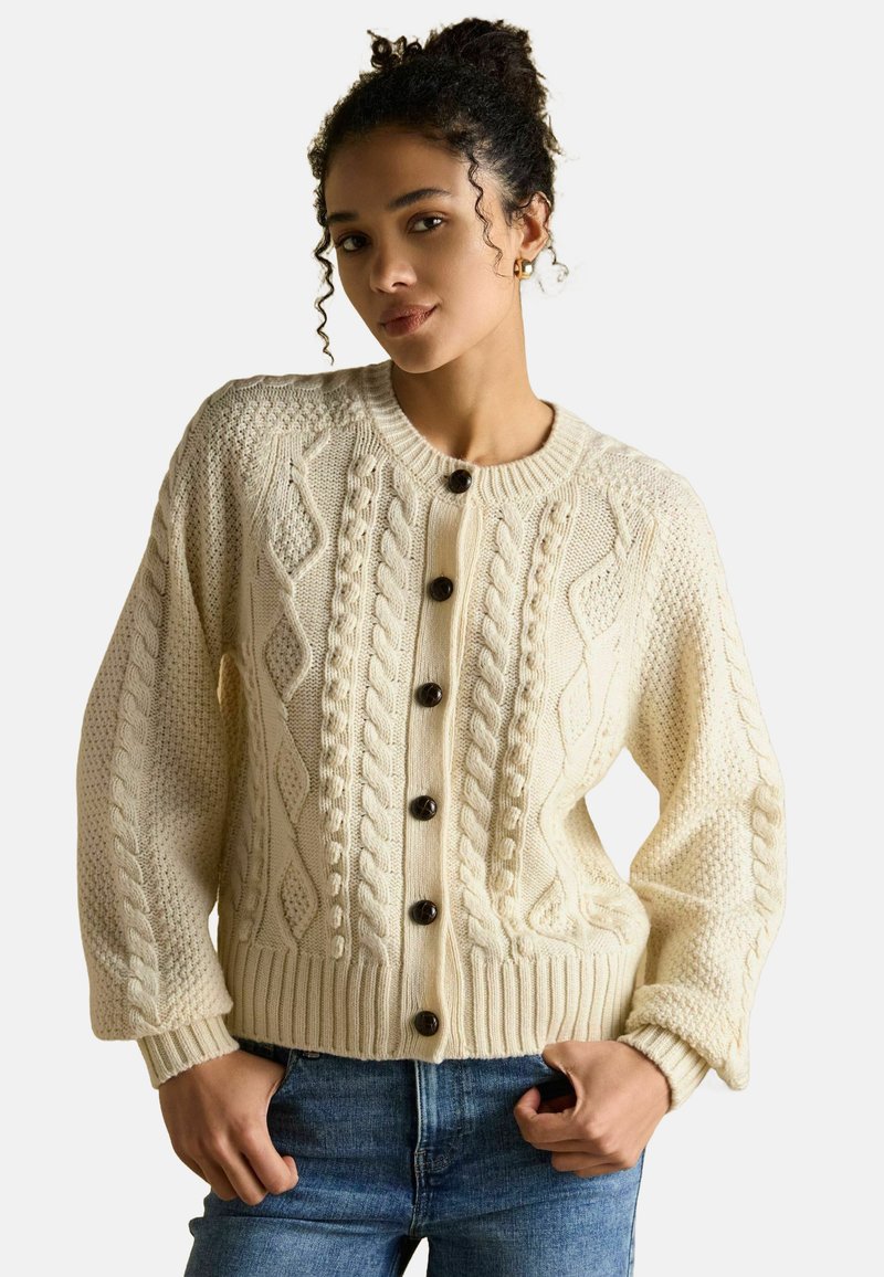 Krémový káblový úpletový sveter s rebrovanými manžetami a lemom, s veľkými tmavými gombíkmi a textúrovanými vzormi, nosený cez modré džínsy.