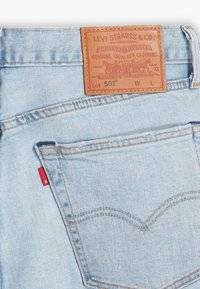 Ljusblå denimjeans med en brun läderlapp med präglad text och en röd märkesflik på bakfickan. Klassisk sömnadsmodell.