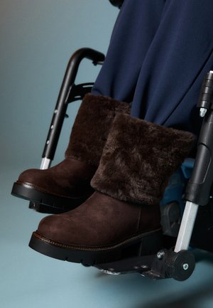 Cilvēks, kurš valkā tumši zilus bikses un brūnus zābakus ar kažokādas oderi, sēž ratiņkrēslā ar pēdām atpūtušām uz kāju balsta.