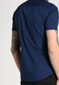 Uomo che indossa una camicia a maniche corte blu navy, con tatuaggi su entrambi i avambracci, in piedi con la schiena rivolta verso lo spettatore.