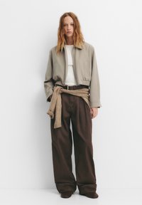 Beige cropped jakke over en hvid top, parret med oversized brune bukser og et brunt bælte. Sweater bundet om livet, glat tekstur.