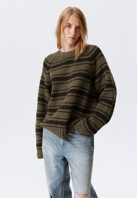 Olivengrønn og mørkebrun stripete strikkegenser med avslappet passform, ribbestrikket halslinning og mansjetter, sammen med slitte lyseblå jeans.