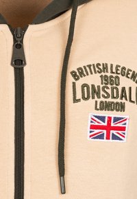 Béžová mikina na zips s tmavozelenými akcentmi, s vyšívaným textom a nášivkou s britskou vlajkou. Predný zips a šnúrka na kapucni.