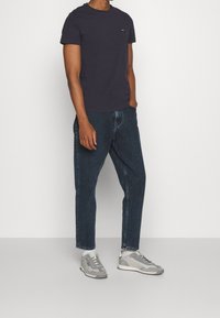 Man som bär svart t-shirt med korta ärmar, mörkblå jeans, vita strumpor och grå sneakers, stående mot en enkel ljus bakgrund.