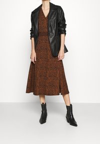 Frau trägt ein braun gemustertes Midikleid, eine schwarze Lederblazerjacke und schwarze Ankle Boots und steht vor einem schlichten weißen Hintergrund.