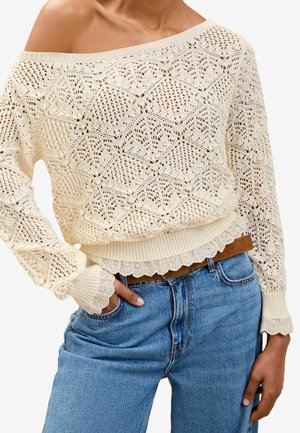 Kvinde iført off-shoulder creme striktrøje med diamantmønster, bølgede kanter, blå jeans og brun bælte, hånd i lommen.