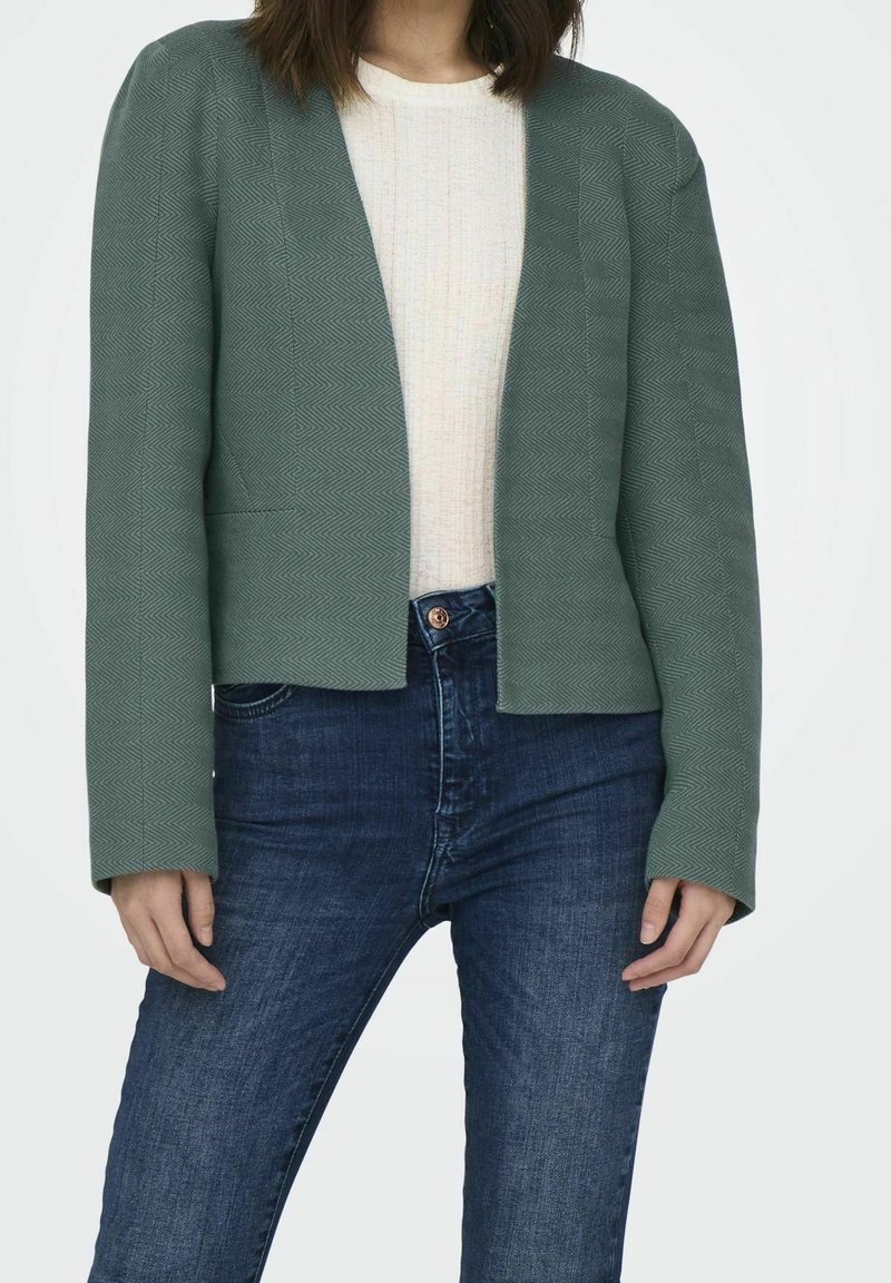 Blazer vert texturé à devant ouvert, manches longues et coupe ajustée, porté sur un haut clair et associé à un jean foncé.
