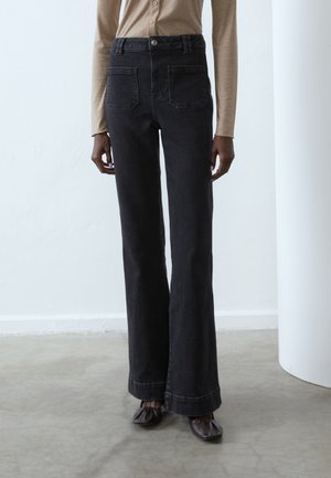 Personne portant un jean noir taille haute à jambes larges avec poches plaquées devant, un haut beige à manches longues et des chaussures carrées marron foncé.
