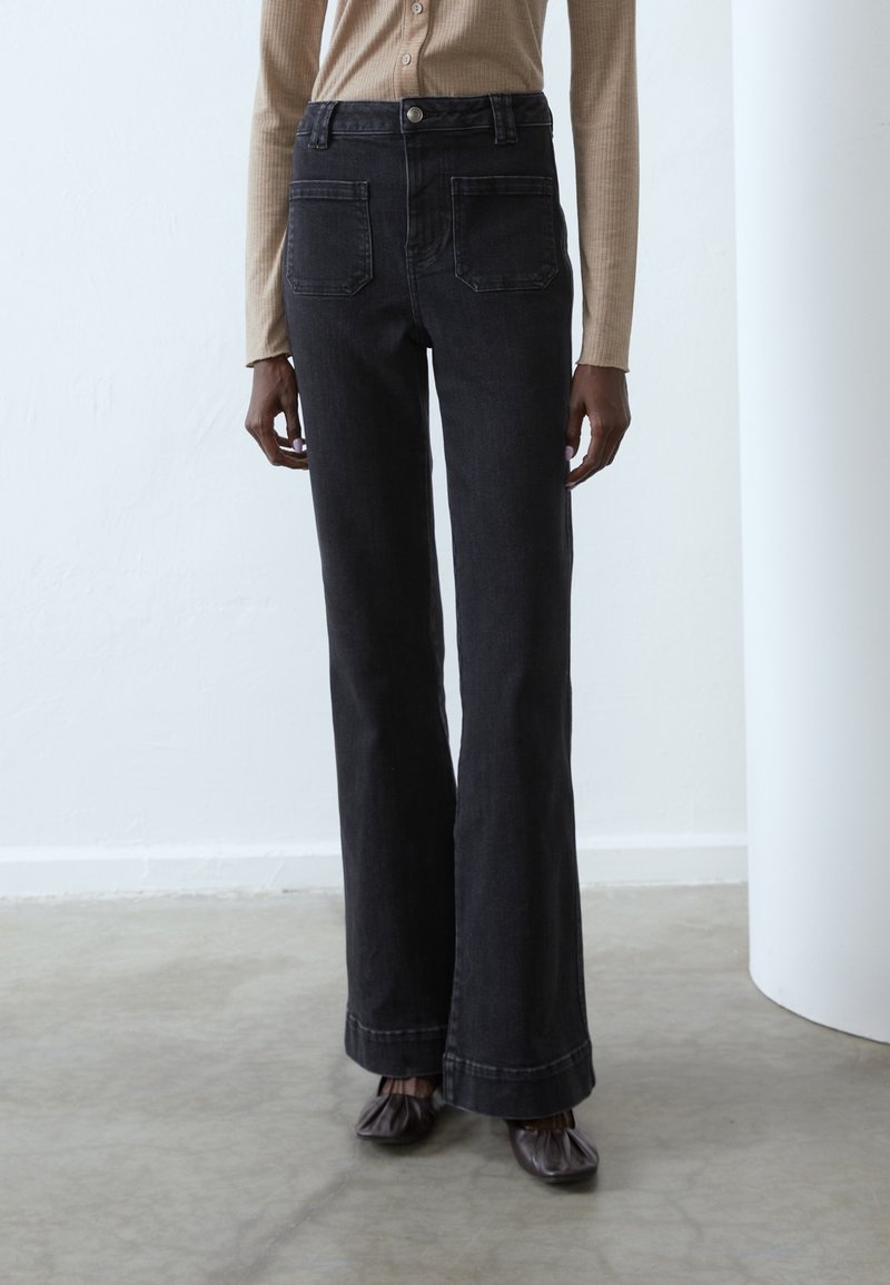 Personne portant un jean noir taille haute à jambes larges avec poches plaquées devant, un haut beige à manches longues et des chaussures carrées marron foncé.