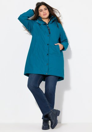 Teal waterproof coat with a hood, front button closure, two side pockets, and a slightly longer back. Paired with blue jeans and black boots.