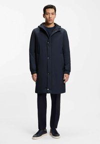 Hombre de pie con un abrigo largo azul marino con botones, pantalones negros y zapatillas negras con suelas blancas, sobre un fondo blanco liso.