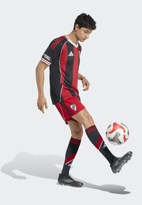 Equipación de fútbol negra y roja con acentos blancos, con mangas cortas y medias a rayas. Jugador conduciendo un balón. Botas negras.