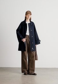 Πολύ μεγάλο μπλε μπουφάν με μεγάλο γιακά και κουμπιά μπροστά, φορεμένο πάνω από λευκό τοπ και καφέ παντελόνια με φαρδιά μπατζάκια. Περιλαμβάνει καφέ τσάντα και καπέλο.