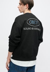 Črna majica s kapuco v prevelikem kroju, z belim in modrim vezenim napisom "OBEY" in "SOUND & RESISTANCE" na hrbtu.