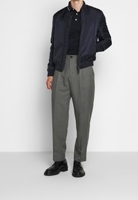 Marinblå bomberjacka över en svart texturerad polo, kombinerad med ljusgrå veckade byxor och svarta läderskor med en slät yta.