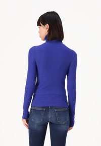 Kuningan berkerah tinggi berwarna biru royal dengan rib yang memiliki lengan panjang; dilengkapi dengan aksen kancing di pergelangan tangan. Dipadukan dengan celana jeans biru yang pas.