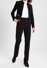 Žena v černé zipové bundě, bílé košili, černých kalhotách s červenými bočními pruhy a černých botách s špičatou špičkou, stojí s jednou rukou v kapse.