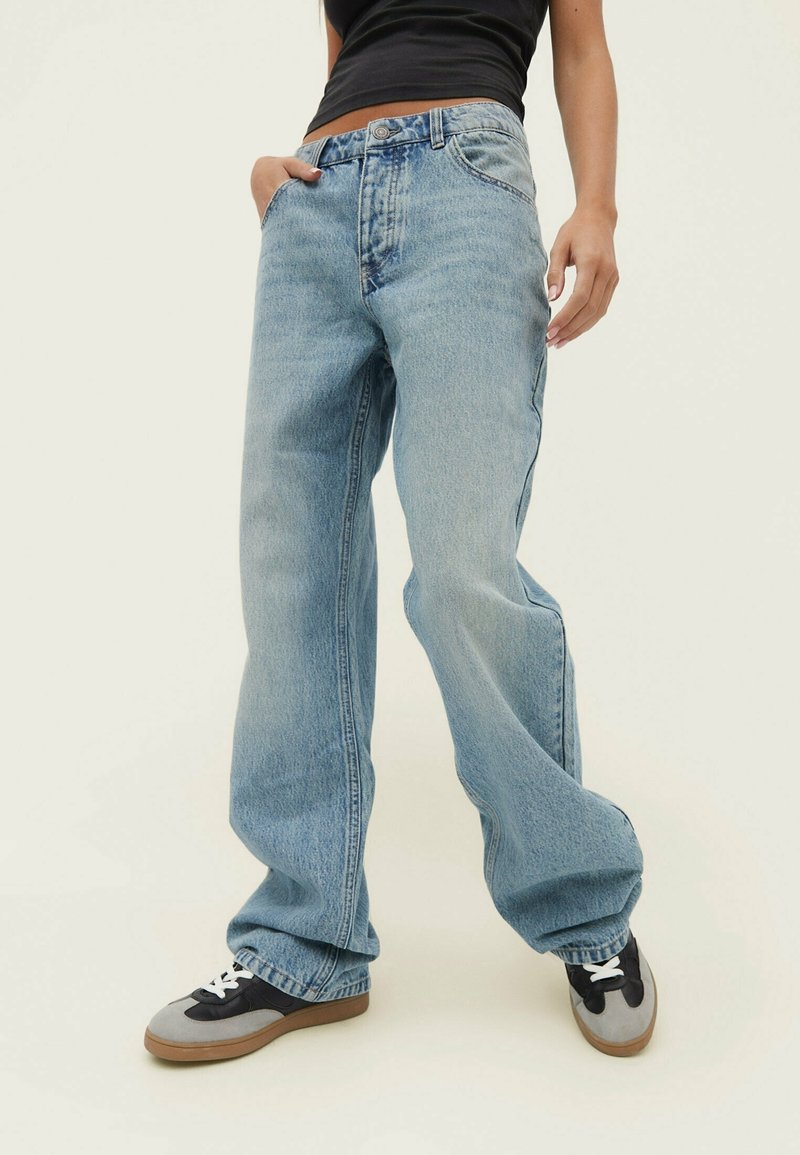 Jeans in denim celeste con vestibilità ampia, dotati di zip frontale, design a cinque tasche e orlo sfilacciato. Abbinati a sneaker nere e grigie.