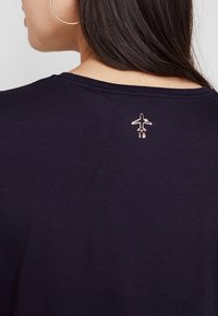 Námornícky tričko s metalickým designem letadla nad zadním výstřihem. Měkká tkanina s volným střihem a jednoduchým kulatým výstřihem.