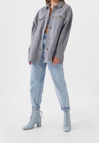 Persoon in lichtblauwe high-waist jeans, chunky lichtblauwe veterschoenen tot de enkel, grijze oversized knoopjas en een cropped top.