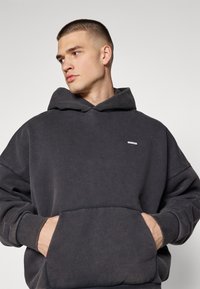 Μαύρη φούτερ ζακέτα με μπροστινή τσέπη, ριμπ μανσέτες και ένα μικρό λογότυπο στο στήθος, κατασκευασμένη από μαλακό ύφασμα για άνεση.