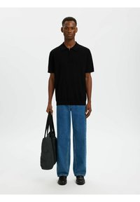 Jeune homme debout tenant un sac fourre-tout noir, portant un polo noir à manches courtes, un jean bleu et des chaussures noires, devant un fond uni.