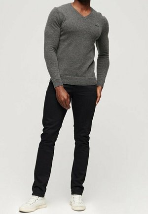 Grauer, gestrickter V-Ausschnitt-Pullover kombiniert mit schwarzen, schmal geschnittenen Jeans und weißen Sneakers. Der Pullover hat gerippte Bündchen und einen gerippten Saum.