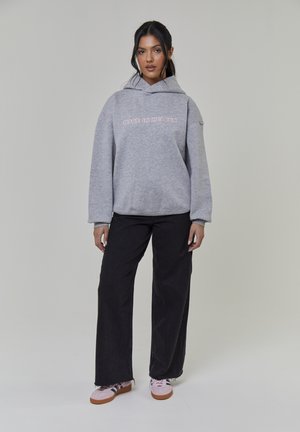 Grauer Kapuzenpullover mit dem Schriftzug "MADE IN HEAVEN", oversized Schnitt, und schwarze Schlagjeans. Die Schuhe sind pinke Sneaker mit einem strukturierten Design.